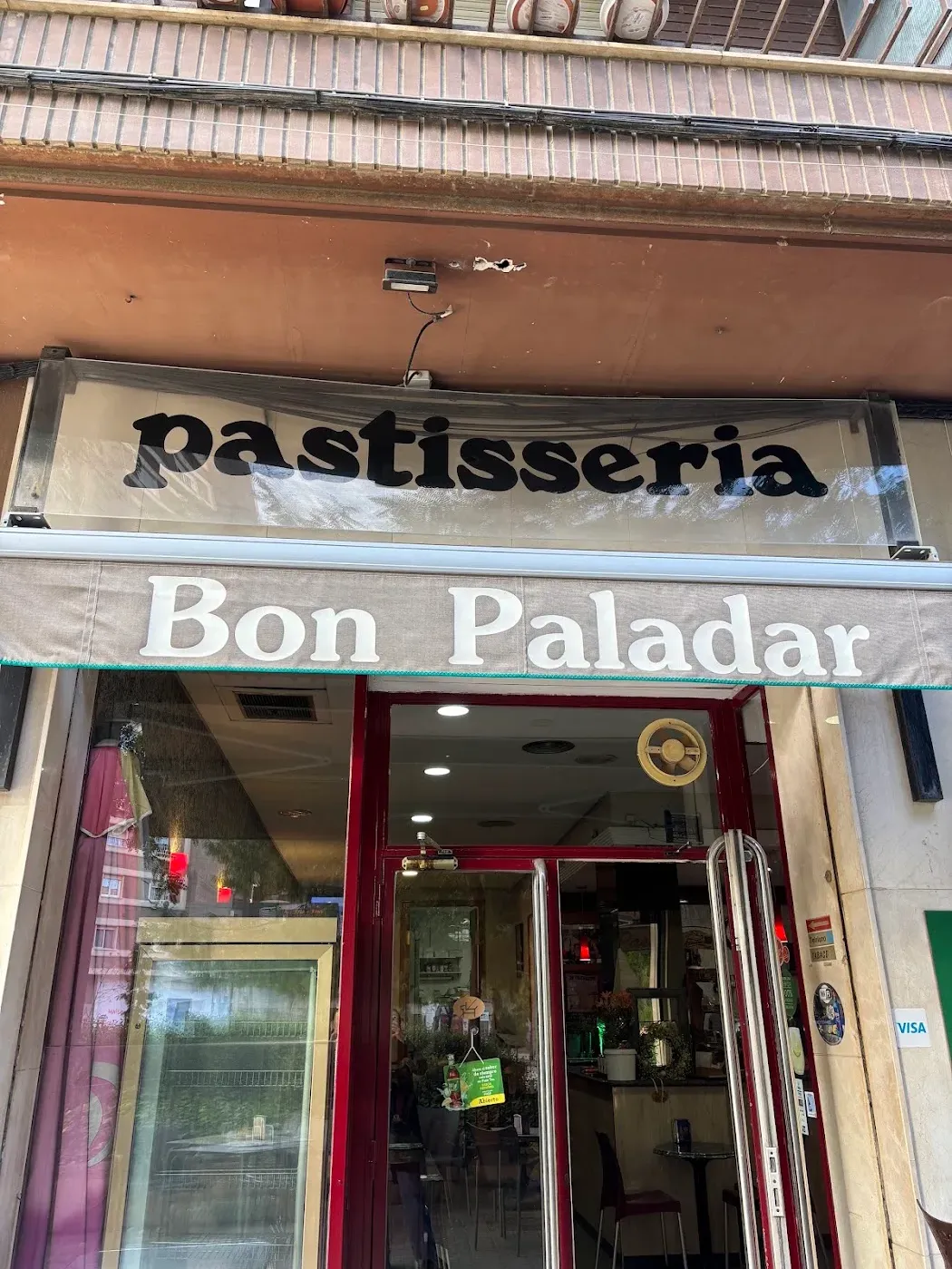 Fachada de Bon Paladar: el rótulo dice «pastisseria» en letras negras sobre el toldo crema y debajo «Bon Paladar» en blanco, con la puerta granate abierta.
